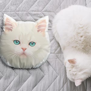 May include: A white cat-shaped pillow with blue eyes and a pink nose rests on a gray, quilted surface. Beside it, a fluffy white cat is curled up. The pillow is a decorative item, and the cat is a pet.
