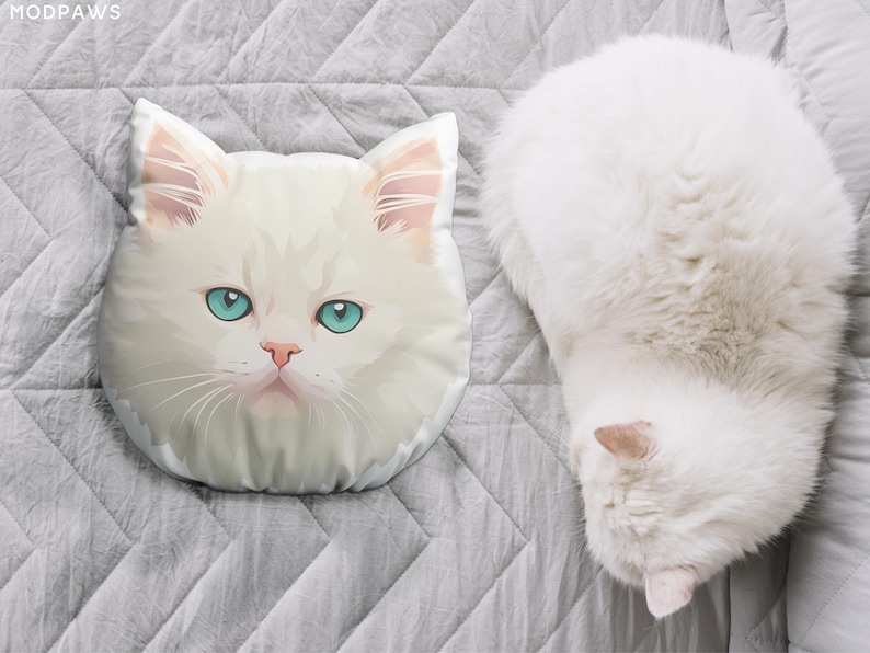 May include: A white cat-shaped pillow with blue eyes and a pink nose rests on a gray, quilted surface. Beside it, a fluffy white cat is curled up. The pillow is a decorative item, and the cat is a pet.