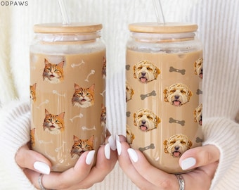 May include: Two clear glass tumblers with bamboo lids and glass straws, filled with light brown iced coffee. The left glass features a repeating pattern of orange tabby cat faces and white fish bones. The right glass shows golden dog faces and bones.