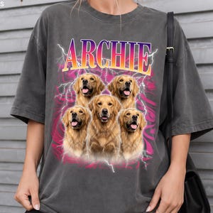 May include: A charcoal gray short-sleeve t-shirt featuring a graphic print of five golden retriever dogs. Above the dogs, the name ARCHIE is printed in a yellow-to-purple gradient font. The design includes pink lightning bolts and abstract patterns.