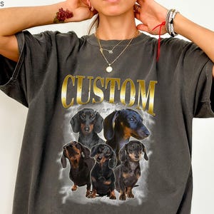 Könnte beinhalten: Ein anthrazitfarbenes Kurzarm-T-Shirt mit dem Wort CUSTOM in großen goldenen Buchstaben über einer grafischen Collage aus fünf schwarz-braunen Dackeln. Das Design hat eine verblasste, vintage-inspirierte Ästhetik mit einem hellgrauen, wolkigen Hintergrund.