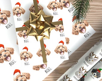 Op de afbeelding: Wit inpakpapier met een herhalend patroon van bruine hondengezichten met rode kerstmutsen en de tekst NOEL onder elk gezicht. Een grote gouden metalen strik is in het midden geplaatst, geaccentueerd door een groene dennentak.