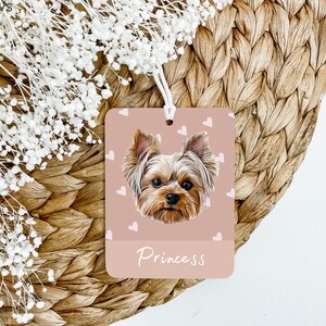 Peut inclure: Un diffuseur de parfum de voiture rose avec une ficelle blanche suspendue en haut. Le diffuseur pr&eacute;sente un Yorkshire Terrier avec une fourrure brune et un fond rose avec des c&oelig;urs blancs. Le texte "Princess" est imprim&eacute; en blanc au bas du diffuseur.