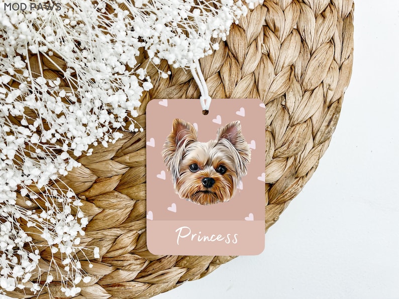 Peut inclure: Un diffuseur de parfum de voiture rose avec une ficelle blanche suspendue en haut. Le diffuseur pr&eacute;sente un Yorkshire Terrier avec une fourrure brune et un fond rose avec des c&oelig;urs blancs. Le texte "Princess" est imprim&eacute; en blanc au bas du diffuseur.
