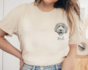 May include: A beige short-sleeve t-shirt featuring a black line art illustration of a dog's face on the left chest area. Below the dog illustration, the name Milo is printed in a black cursive script. The text MODPAWS appears in the top left corner.