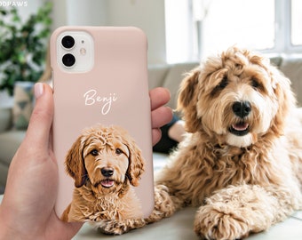 Op de afbeelding: Een zachtroze telefoonhoesje met een portret van een Goldendoodle-hond en de naam "Benji" in cursief schrift. De afbeelding van de hond heeft bruine en beige vacht. De telefoonhoes wordt in een hand gehouden, met een Goldendoodle-hond op de achtergrond.