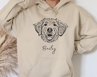 Puede incluir: Una sudadera con capucha de color beige que presenta una ilustración de línea negra de la cara de un perro centrada en el pecho. Debajo de la ilustración del perro, el nombre Bailey está impreso en cursiva negra. Incluye un bolsillo canguro.