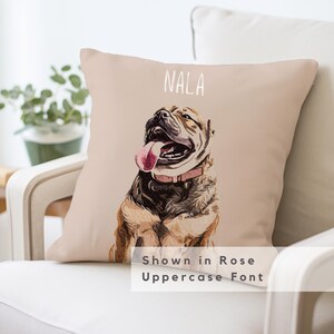 May include: A beige throw pillow with a watercolor illustration of a brown and white dog with a pink collar and tongue sticking out. The dog's name "NALA" is written in white text above the dog. The text "Shown in Rose Uppercase Font" is written below the dog.