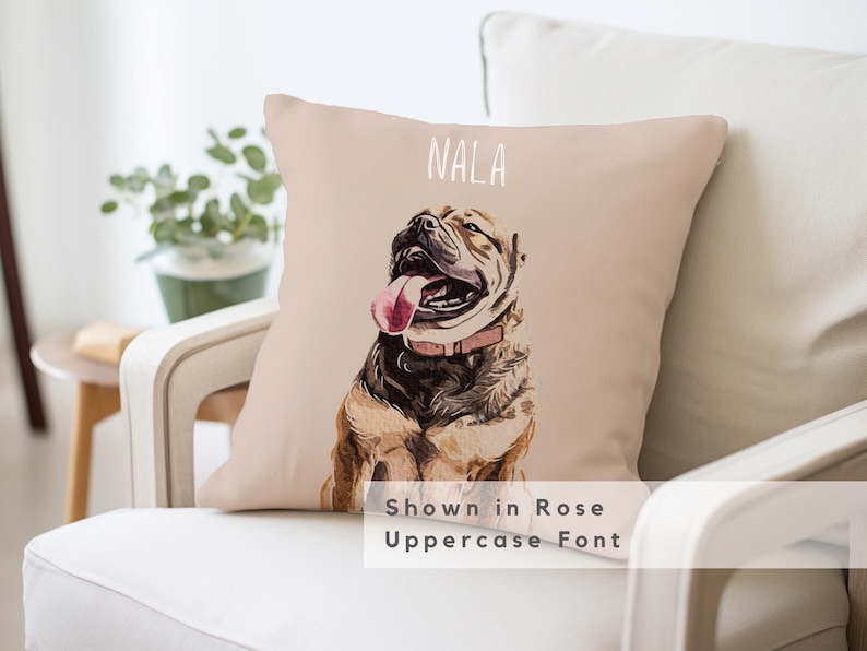 May include: A beige throw pillow with a watercolor illustration of a brown and white dog with a pink collar and tongue sticking out. The dog's name "NALA" is written in white text above the dog. The text "Shown in Rose Uppercase Font" is written below the dog.