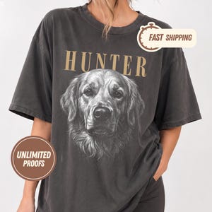 May include: A charcoal grey short-sleeve t-shirt featuring a detailed black and white illustration of a golden retriever dog centred on the chest. Above the dog, the word HUNTER is printed in large, tan, serif capital letters.