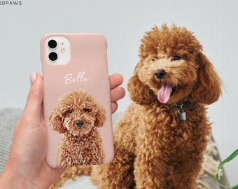Op de afbeelding: Een zachtroze telefoonhoesje met een bruin poedelportret en de naam "Bella" in witte letters. Op de achtergrond is een levende bruine poedel te zien. De telefoonhoes wordt in een hand gehouden.
