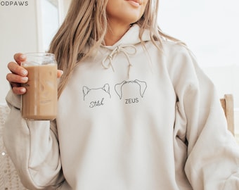 Puede incluir: Una sudadera con capucha de color beige que incluye un cordón ajustable y un gráfico de líneas negras en el pecho. El diseño muestra dos pares de orejas de animal con los nombres Stitch y ZEUS impresos en negro debajo de cada par.