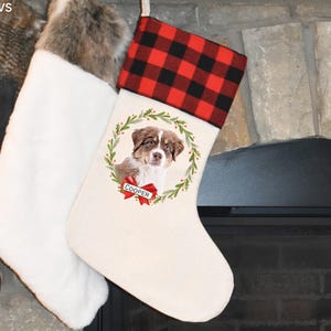 Puede incluir: Una media de Navidad blanca con un puño de cuadros rojos y negros. La media tiene una corona con un perro marrón y blanco dentro y el nombre "Cooper" en una cinta roja.