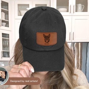 May include: Black baseball cap with a brown leather patch featuring a dog illustration and the name "Winston". The cap has a curved brim and a classic, casual style. The text "Designed by real artists!" is at the bottom.
