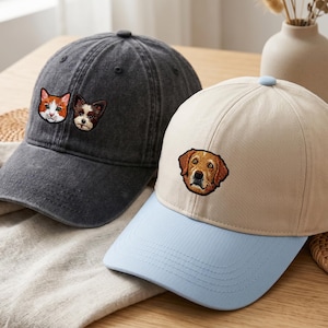 以下が含まれることがあります： オレンジと白の猫と小さな茶色の犬が刺繍されたチャコールグレーの野球帽と、ライトブルーのつばが付いたベージュの野球帽。ベージュの帽子のフロントパネルには茶色の犬の顔が刺繍されています。