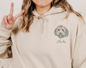 Puede incluir: Una sudadera con capucha de color beige que presenta una ilustración de línea negra de la cara de un perro en el lado izquierdo del pecho. Debajo de la ilustración del perro, el nombre Charlie está impreso en una fuente cursiva negra.