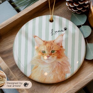 Pode incluir: Enfeite redondo de cer&acirc;mica com um retrato em aquarela de um gato laranja com olhos verdes, chamado Luna. O fundo tem listras verticais verdes e brancas. Uma corda dourada &eacute; anexada para pendurar. O enfeite &eacute; exibido em uma bandeja de madeira.