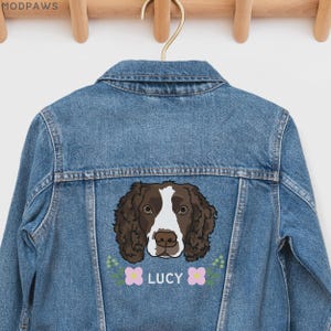 May include: A blue denim jacket featuring a graphic illustration of a brown and white spaniel dog on the back. Below the dog, the name LUCY is printed in white capital letters, flanked by two small pink flowers and green leaf accents.