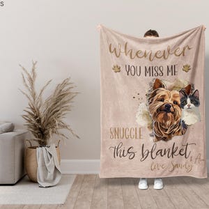 May include: A beige blanket with the text "Whenever you miss me, snuggle this blanket." The blanket features illustrations of a dog and a cat. The blanket is held up by a person wearing white shoes.