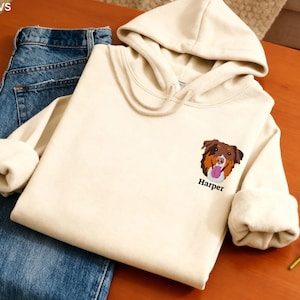 Può includere: Felpa con cappuccio color crema con un ricamo di un muso di cane marrone e beige sul davanti, con il nome "Harper" sotto. La felpa con cappuccio è piegata accanto a un paio di jeans blu. L'immagine è su una superficie di legno.
