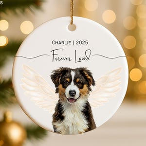May include: A white ceramic Christmas ornament with a gold hanging string. The ornament features a color illustration of a dog with angel wings, the name "Charlie | 2025", and the words "Forever Loved".