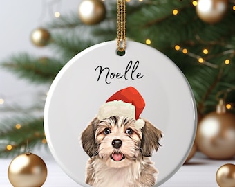 May include: A round white ceramic ornament featuring a digital illustration of a fluffy brown and white puppy wearing a red and white Santa hat. The name Noelle is printed in black script above the puppy. A gold string is attached to the top.