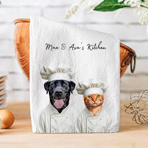 Op de afbeelding: Een witte keukendoek met een grafische print van een zwarte Labrador-hond en een oranje cyperse kat die witte koksmutsen en witte dubbelrijige koksbuizen dragen. De tekst Max & Ava's Kitchen staat in zwarte letters boven de dieren gedrukt.