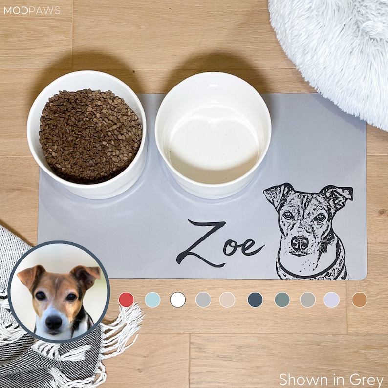 May include: A grey pet feeding mat with two white ceramic bowls. The mat has a dog portrait and the name "Zoe" in black. The text "Shown in Grey" is at the bottom. A small dog portrait is in the corner.