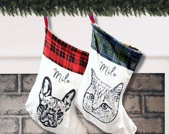 May include: Two white Christmas stockings featuring black ink illustrations of a French Bulldog and a cat. The left stocking has a red and black plaid cuff, while the right has a green and blue plaid cuff. Both stockings feature the name Milo.