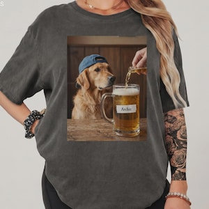 Può includere: Una t-shirt a maniche corte grigio antracite con una stampa grafica rettangolare di un golden retriever che indossa un berretto blu, seduto a un tavolo di legno accanto a un boccale di birra con il nome Archie su un'etichetta bianca.