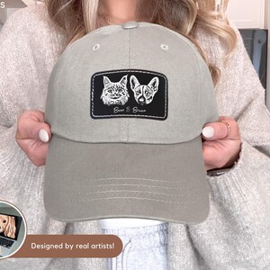 May include: A light gray baseball cap with a patch featuring illustrations of a cat and a dog, with the text "Bear & Bruno". The cap has a curved brim and a button on top. The text "Designed by real artists!" is also visible.