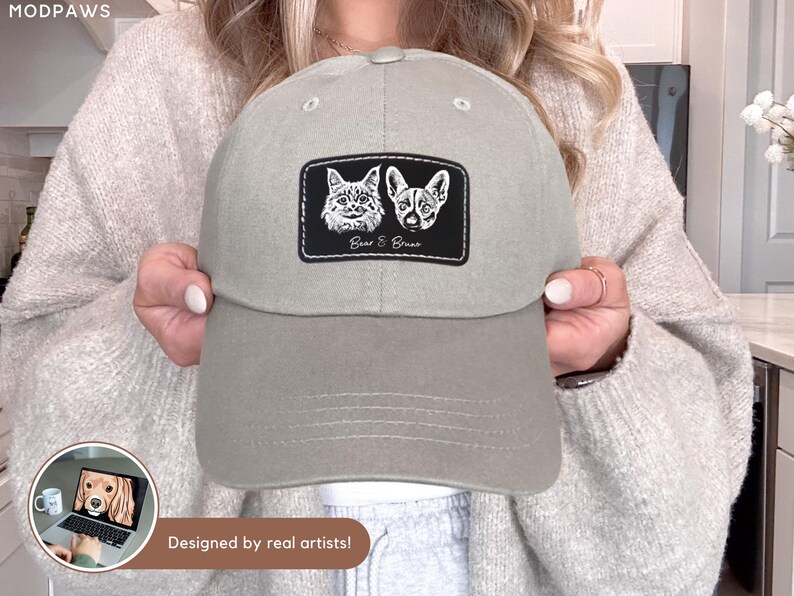 May include: A light gray baseball cap with a patch featuring illustrations of a cat and a dog, with the text "Bear & Bruno". The cap has a curved brim and a button on top. The text "Designed by real artists!" is also visible.