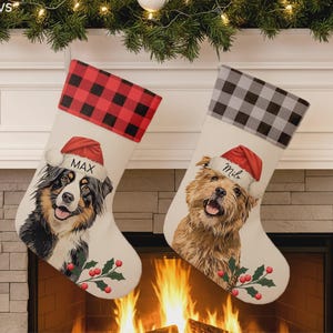 Puede incluir: Dos calcetines navideños de color blanco roto con ilustraciones de perros con gorros de Papá Noel. El calcetín izquierdo tiene un puño de cuadros rojos y negros y el nombre MAX. El derecho tiene un puño de cuadros grises y negros y el nombre Milo.