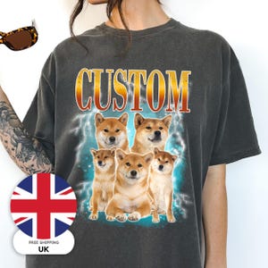 May include: Dark grey t-shirt with a graphic of five Shiba Inu dogs, set against a blue lightning effect. The word "CUSTOM" is printed in a gold and orange gradient font above the dogs. The shirt has a relaxed fit.