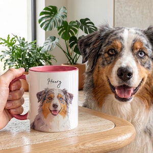 May include: A white ceramic mug with a pink interior and handle, featuring a colorful illustration of an Australian Shepherd dog and the name Henry printed in black script above the dog. The mug is held in a hand next to a dog.