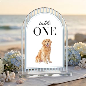 May include: Clear acrylic table number one with a watercolor illustration of a golden retriever. The sign has an arched top and a rectangular base. The words "table ONE" are printed in black.