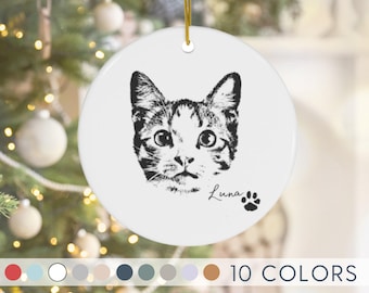 May include: A round white ceramic ornament featuring a black line art illustration of a cat face. The name Luna is printed in a cursive script next to a small black paw print. A gold string is attached to the top for hanging the ornament.