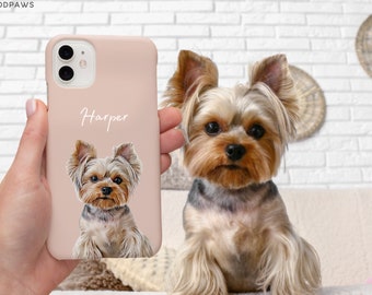 Op de afbeelding: Een zachtroze telefoonhoesje met een portret van een Yorkshire Terrier en de naam "Harper" in cursief. De telefoonhoes wordt vastgehouden, met de hond op de achtergrond. De hond is een klein ras met bruin en bruinachtig haar.