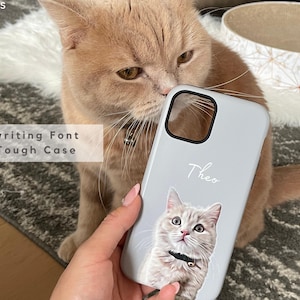 May include: A light gray phone case with a cat portrait and the name "Theo" in handwriting font. The case is a tough case. The image also shows a tan cat.