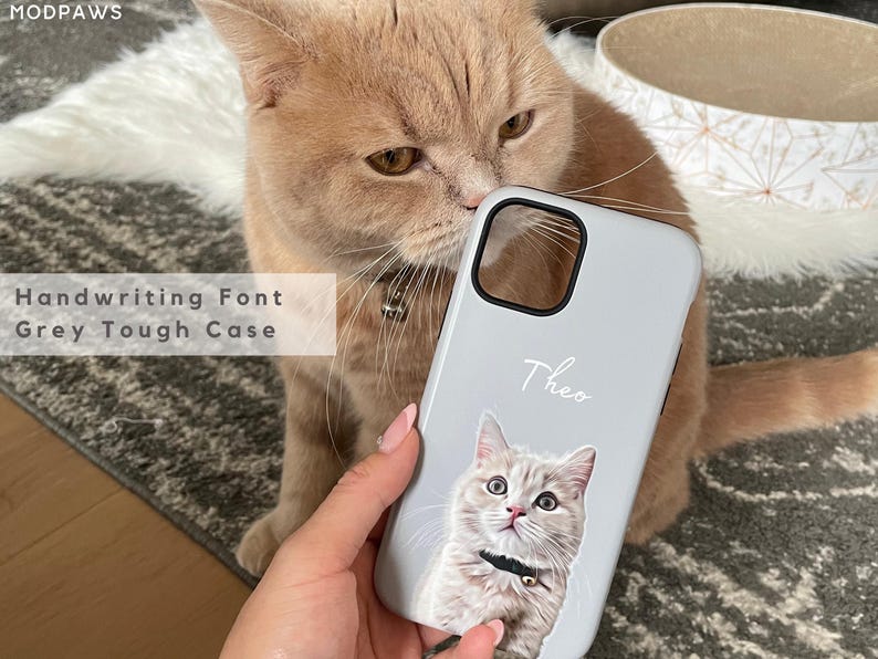 May include: A light gray phone case with a cat portrait and the name "Theo" in handwriting font. The case is a tough case. The image also shows a tan cat.