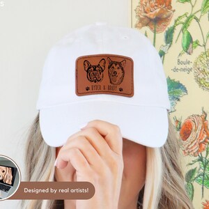 May include: White baseball cap with a brown leather patch featuring two dog portraits and the text "Ryder & Brady". The patch is rectangular and has a paw print on each side of the text. The words "Designed by real artists!" are also visible.
