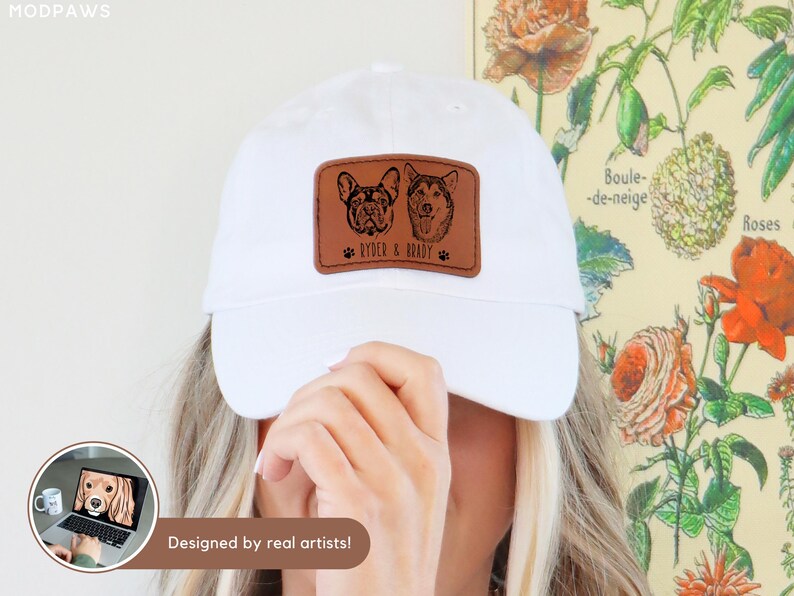 May include: White baseball cap with a brown leather patch featuring two dog portraits and the text "Ryder & Brady". The patch is rectangular and has a paw print on each side of the text. The words "Designed by real artists!" are also visible.