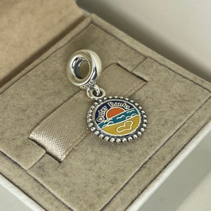 K&ouml;nnte beinhalten: Silberner Anh&auml;nger mit einem blauen, gelben und orangefarbenen Emaille-Design eines Sonnenuntergangs &uuml;ber dem Meer. Der Anh&auml;nger tr&auml;gt die Aufschrift "Outer Banks".