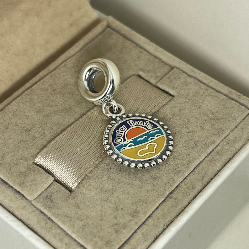 K&ouml;nnte beinhalten: Silberner Anh&auml;nger mit einem blauen, gelben und orangefarbenen Emaille-Design eines Sonnenuntergangs &uuml;ber dem Meer. Der Anh&auml;nger tr&auml;gt die Aufschrift "Outer Banks".