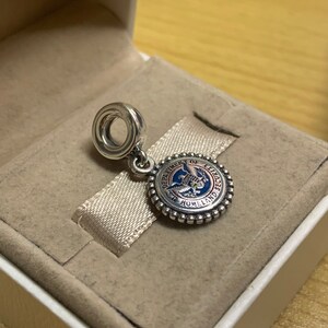 May include: A silver charm with a blue, red, and white enamel design. The charm features the seal of the Department of Homeland Security, which includes an eagle with outstretched wings and the text "Department of Homeland Security".
