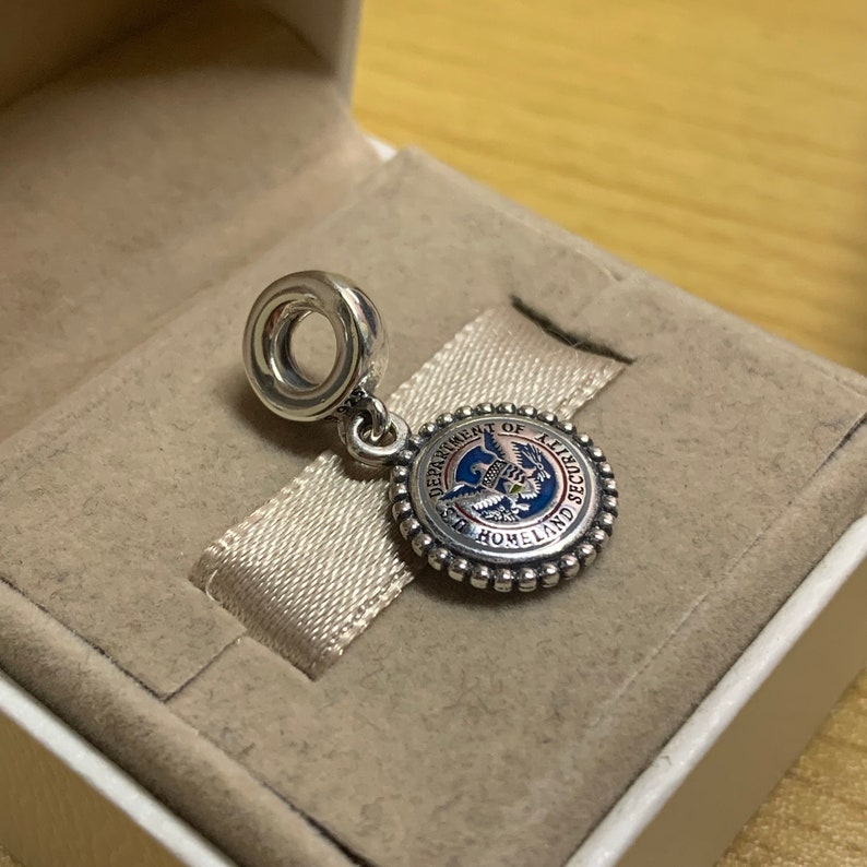 May include: A silver charm with a blue, red, and white enamel design. The charm features the seal of the Department of Homeland Security, which includes an eagle with outstretched wings and the text "Department of Homeland Security".