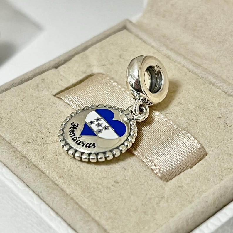Pu&ograve; includere: Un ciondolo d'argento a forma di cuore blu e bianco con un design di bandiera e la parola "Honduras" in lettere bianche.