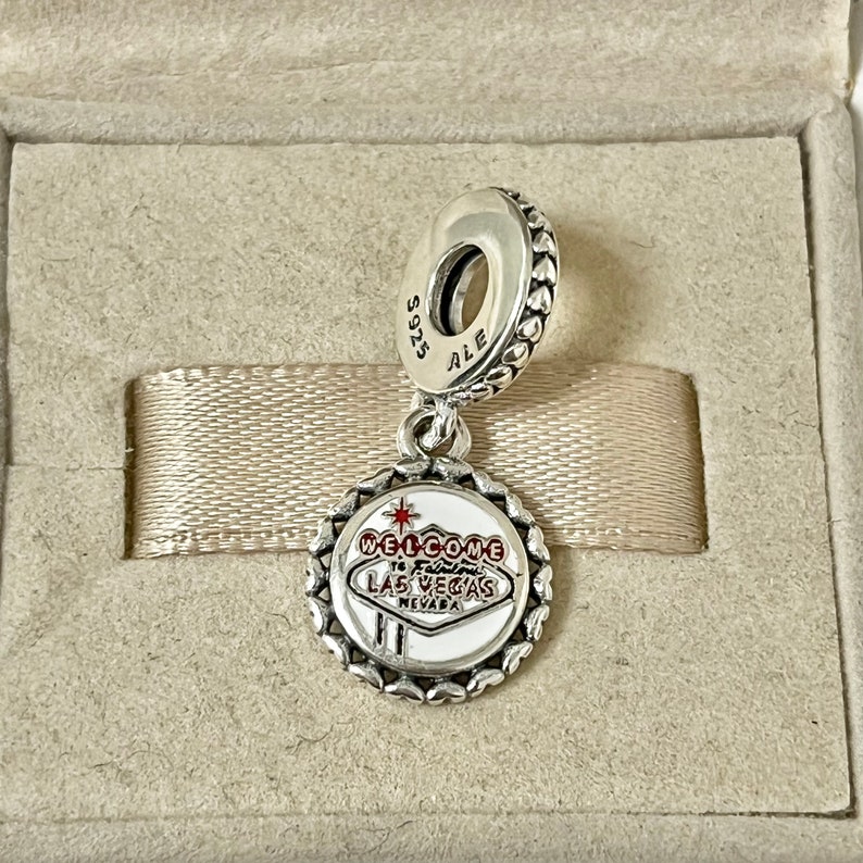 Puede incluir: Encanto de plata con un letrero de esmalte blanco que dice "Welcome to Fabulous Las Vegas Nevada". El encanto tiene un borde de coraz&oacute;n plateado y un lazo plateado en la parte superior.