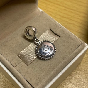 May include: A silver charm with a round, beaded edge and a circular disc in the center. The disc has a raised "O" with a smaller circle in the center and the word "PANDORA" inscribed around the edge.