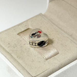 May include: A silver heart-shaped charm with a black and red snake design. The charm has the word "ALE" engraved on it. The charm is in a white box with a beige interior.